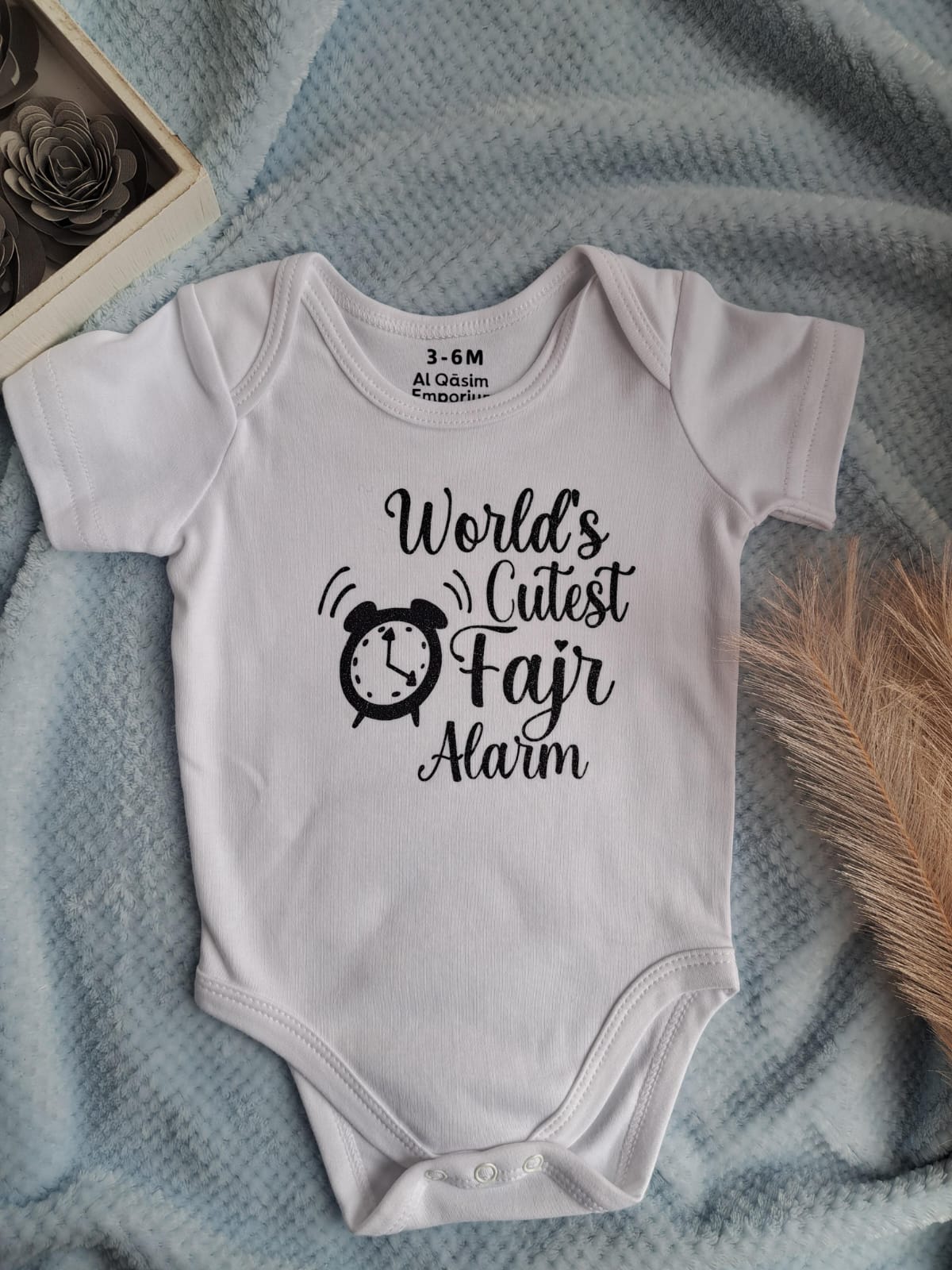 White baby onesie with 'World's Cutest Fajr Alarm' text on a textured surface